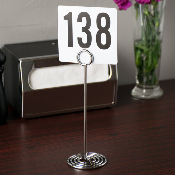 An American Metalcraft chrome swirl base card holder with a number on it on a table with a green plant in a glass.
