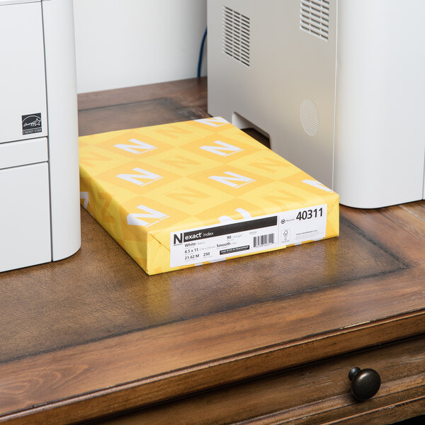 A yellow box of Neenah Exact white cardstock on a desk next to a printer.