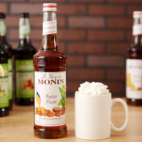 A close-up of a bottle of Monin Butter Pecan flavoring syrup with a white label next to a mug of coffee with whipped cream on top.