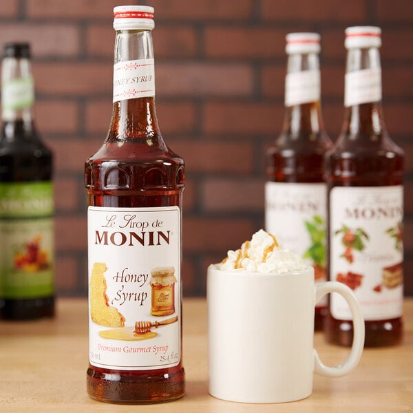 A close-up of a Monin Premium Honey Syrup bottle next to a mug of whipped cream with caramel.