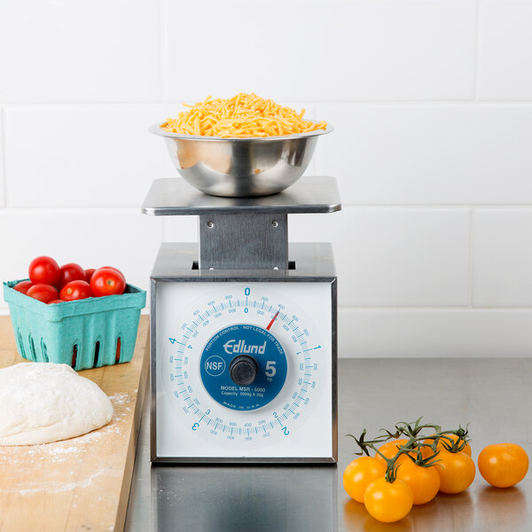 An Edlund stainless steel portion scale on a counter with a bowl of noodles on top and a bowl of tomatoes on the counter.