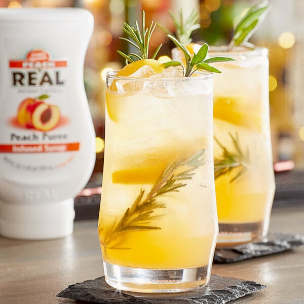 A table with two glasses of peach and lemonade with rosemary sprigs next to a bottle of Real Peach Puree Infused Syrup.