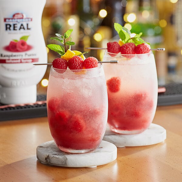 A pair of glasses with red raspberry drinks on a coaster.