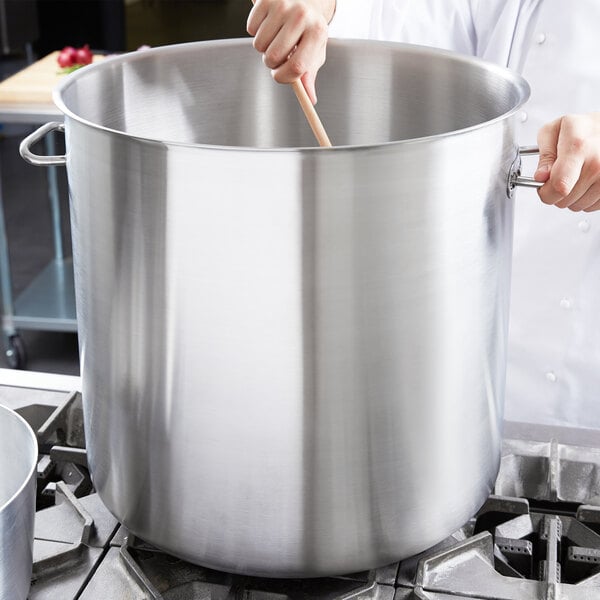 A person stirring a large silver Vollrath stock pot with a wooden spoon.