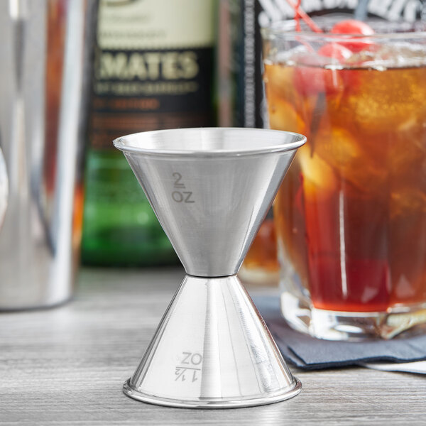 An American Metalcraft stainless steel jigger filled with liquor on a table next to a drink.