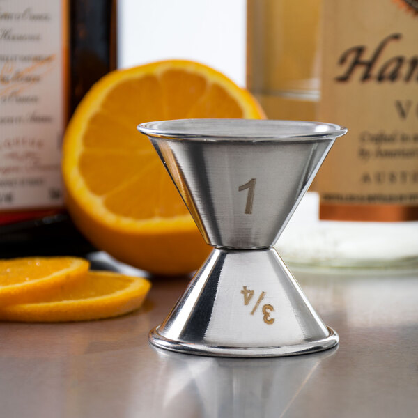 An American Metalcraft stainless steel jigger with orange slices and a bottle of alcohol on a counter.