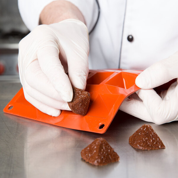 A gloved hand uses a Matfer Bourgeat orange silicone mini pyramid mold to make brown candies.
