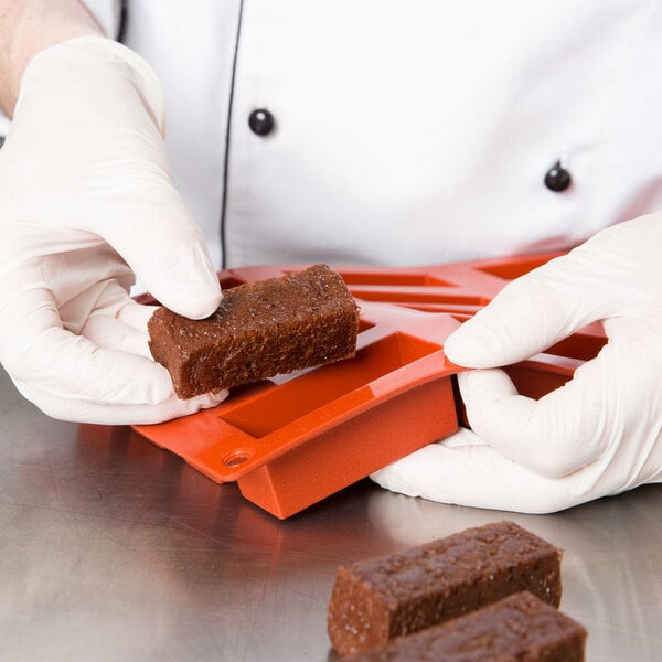 A person's hand using a red Matfer Bourgeat silicone rectangular mold to make mini cakes.
