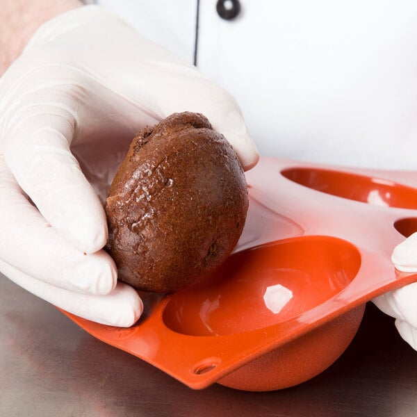A gloved hand using a Matfer Bourgeat orange silicone half sphere mold to hold a brown round object.