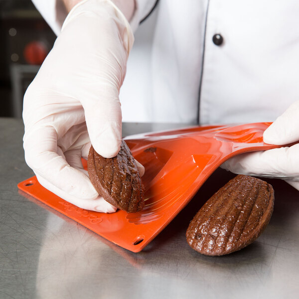 A person in gloves holding a brown Matfer Bourgeat Madeleine cookie.