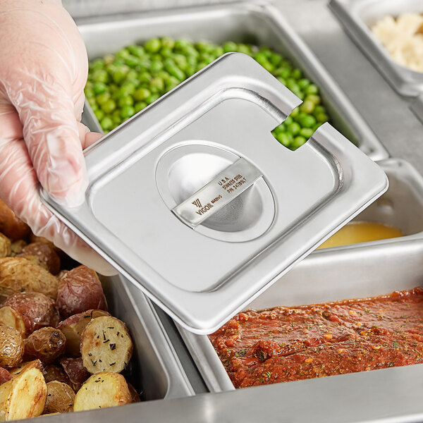 A person holding a Vigor stainless steel pan with a metal lid on a tray of food.