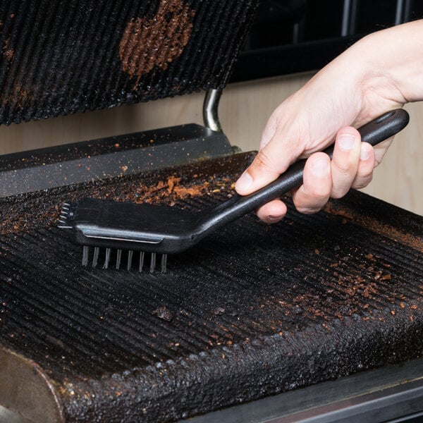 A person using a Chef Master panini grill brush with a black handle to clean a grill.