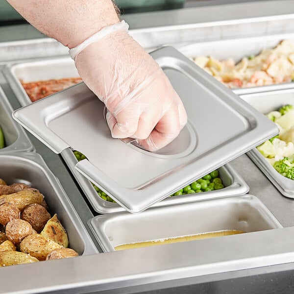 A hand in a plastic glove placing a Vigor stainless steel pan cover on a tray of food.