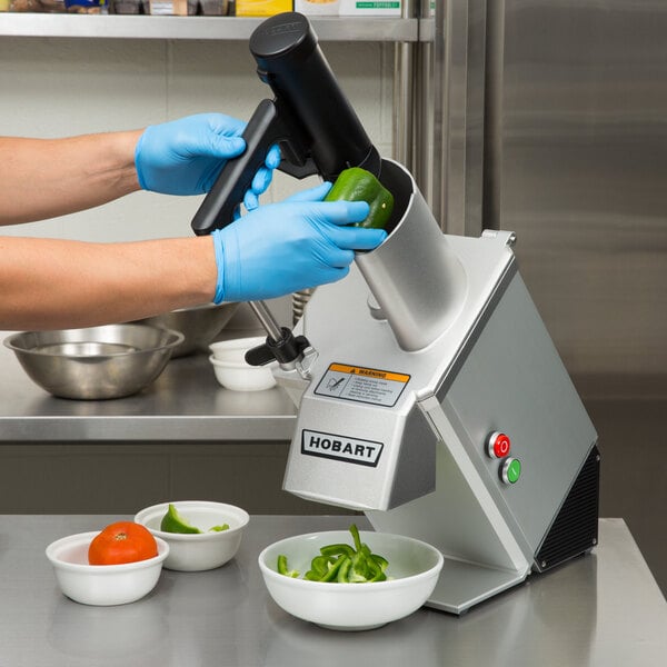 A person in blue gloves using a Hobart food processor to cut green peppers.