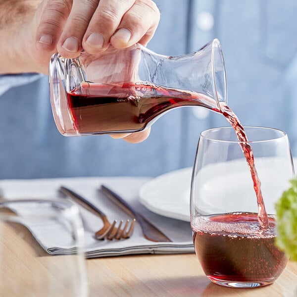 A hand pouring red wine into a clear Acopa glass carafe on a table with a fork and knife.