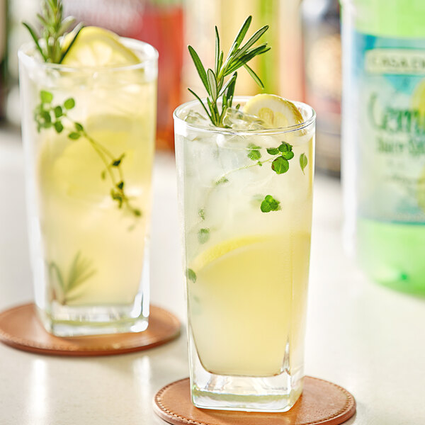A close-up of two glasses of lemonade with rosemary and a bottle of Natural Strength Lemon Juice Splash on a table.