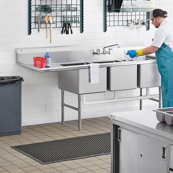 A man in a blue uniform washing dishes in a commercial kitchen using a Regency three compartment sink with a left drainboard.