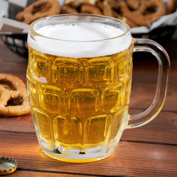 A Libbey beer mug filled with beer with pretzels on a wooden table.