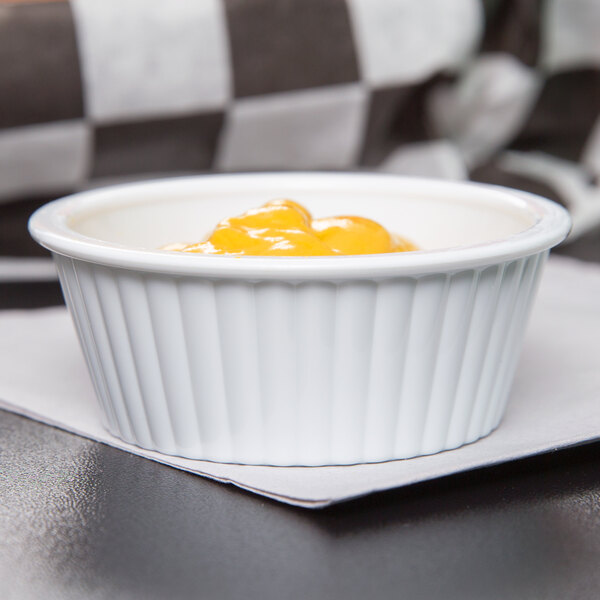 A white fluted plastic GET ramekin filled with yellow liquid on a table with a blurry black and white checkered surface.