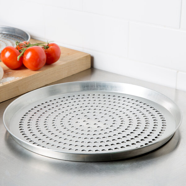 An American Metalcraft tin-plated steel deep dish pizza pan with perforations on a metal surface next to tomatoes.