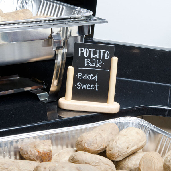 An American Metalcraft natural wood-finish table top board with a sign on a table next to potatoes.