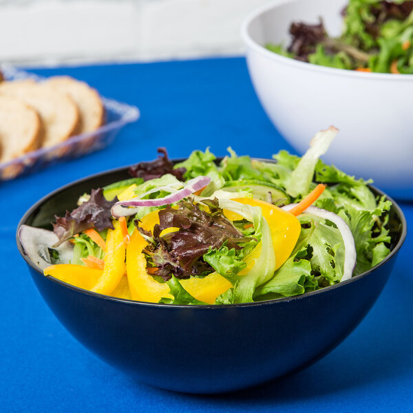 A Fineline black plastic bowl filled with salad on a blue table.