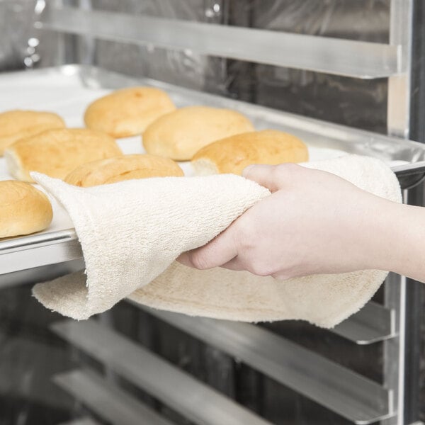 A hand holding a San Jamar terry cloth towel over a tray of baked bread.