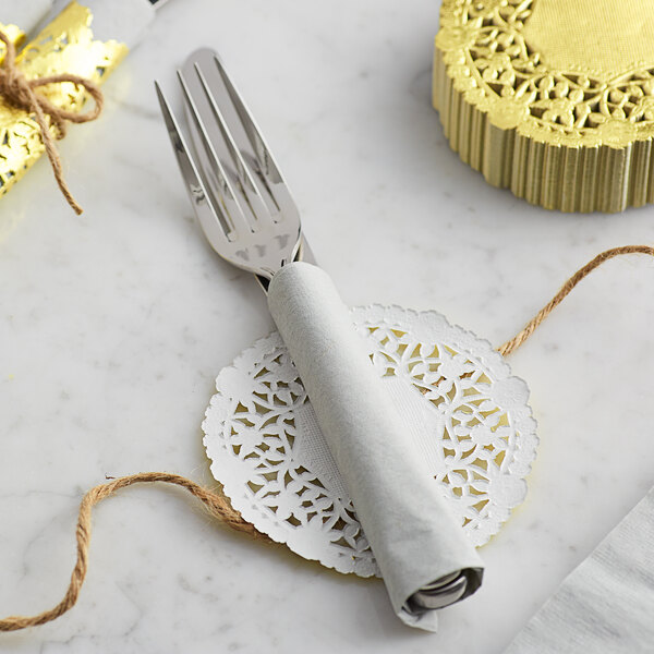 A white table with a fork wrapped in a 4" gold foil lace doily.