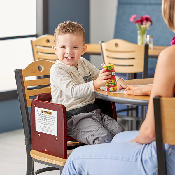 A child sitting in a Lancaster Table & Seating dark mahogany wooden booster seat at a table with a woman holding a juice box.