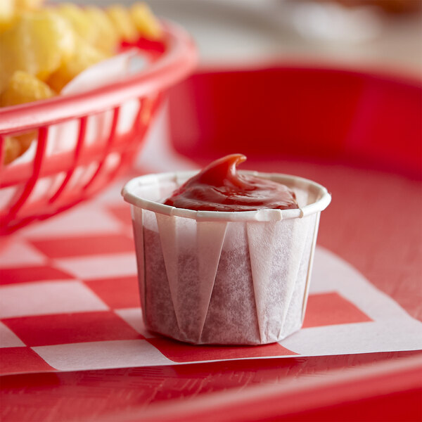 A Genpak Harvest paper souffle cup filled with ketchup on a red and white checkered tablecloth.