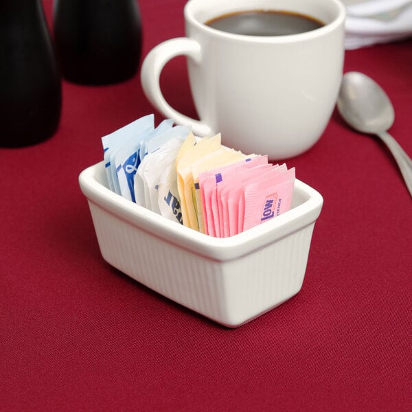 A white Tuxton sugar packet holder with sugar packets next to a cup of coffee.