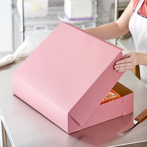 A woman opening a pink Baker's Mark bakery box with a cake inside.