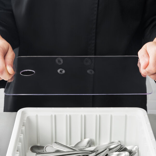 A person holding a clear plastic rectangular object with holes over a tray of spoons and forks.