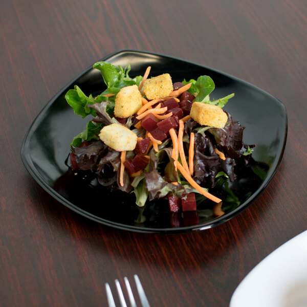 A close-up of a Carlisle black square melamine plate with a salad on a table.