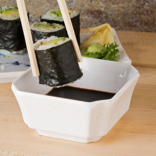 Chopsticks holding a Blue Bamboo square melamine bowl filled with a sushi roll over a bowl of soy sauce.
