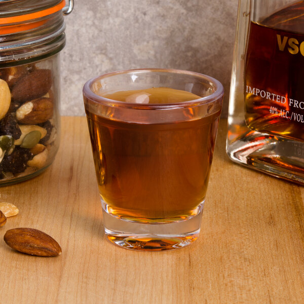 A 1.5 oz. plastic shot glass filled with brown liquid on a table next to a jar of nuts and a bottle of liquor.