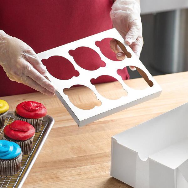 A person holding a white Baker's Mark cupcake box with 6 cupcakes inside.