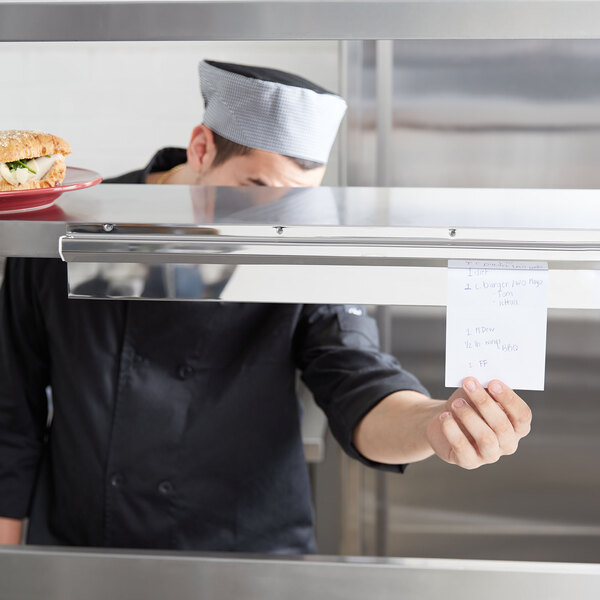 A chef using an Advance Tabco aluminum guest check holder to hold a receipt over a counter with food.