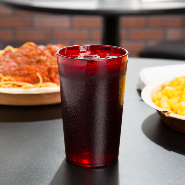 A Cambro ruby red plastic tumbler filled with a dark liquid and ice on a table.