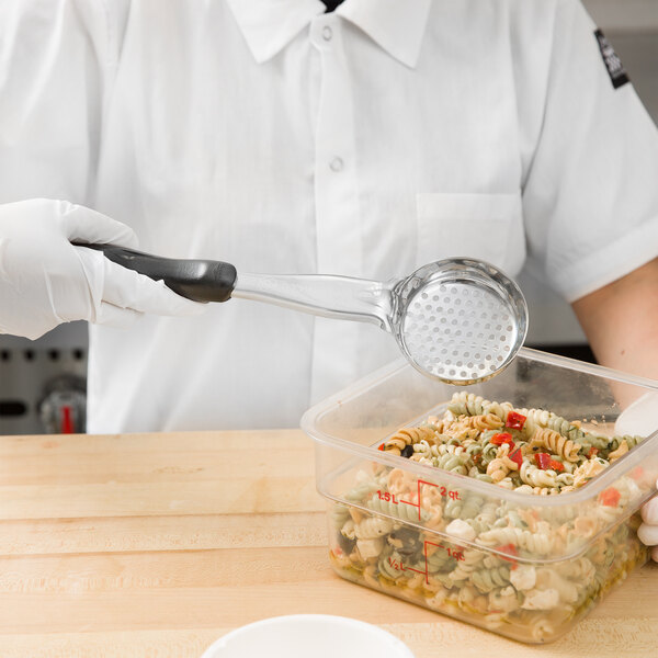 A person in white gloves using a Vollrath black perforated round Spoodle to serve pasta from a plastic container.