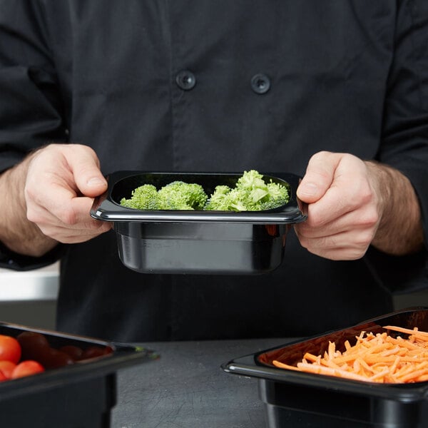 A chef using a Cambro black food pan to serve broccoli and carrots.