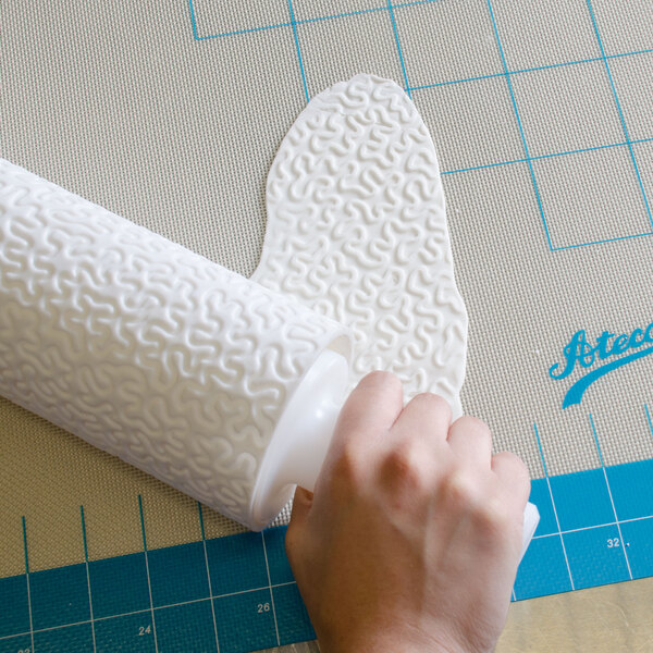 A hand using a white Ateco plastic lace rolling pin cover to roll fondant.