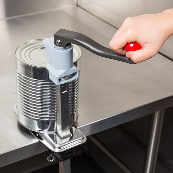 A hand using a Vollrath Redco CanMaster can opener with a red handle to open a can on a table.