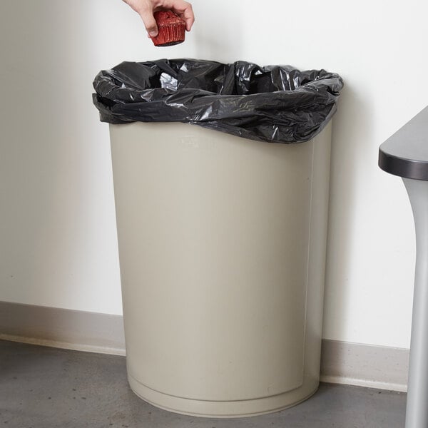 A hand putting a plastic bag into a beige Rubbermaid half round trash can.