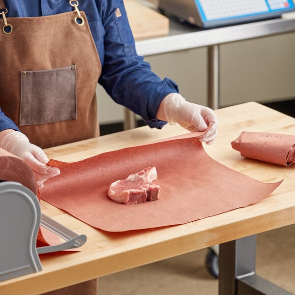A person in a brown apron cutting meat on a counter using Choice pink butcher paper.