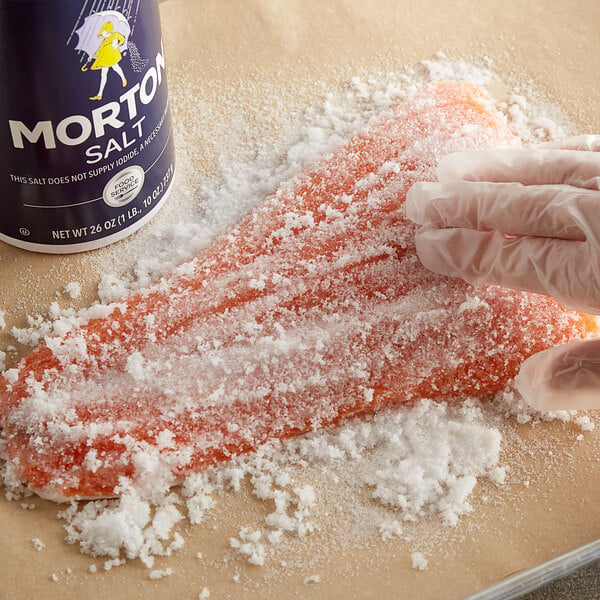 A gloved hand sprinkling Morton plain table salt rounds over a piece of salmon.