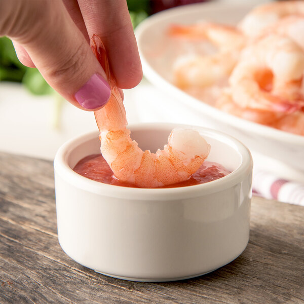A hand holding a shrimp in a Carlisle ivory ramekin of sauce.