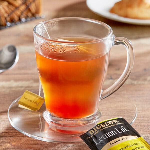 A glass cup of Bigelow Lemon Lift tea with a tea bag on a saucer.