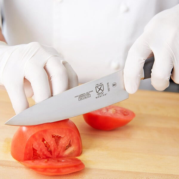 A person wearing white gloves using a Mercer Culinary Genesis Chef Knife to cut a tomato.