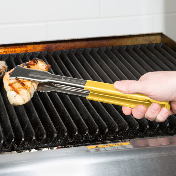 A hand holding yellow Vollrath VersaGrip tongs over a grill.